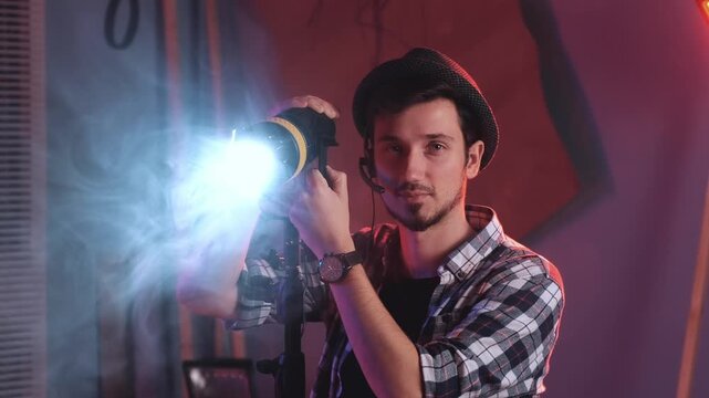 A male technician wearing a plaid shirt and cap adjusts a large lighting fixture in a red-lit film studio filled with smoke and haze.