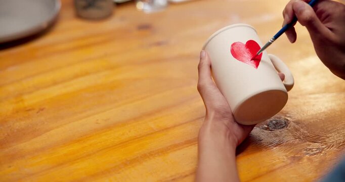 Artisan, painting and cup in hands with space, creative and drawing heart shape with paintbrush. Pottery, craftsmanship and person with handmade mug, above and art process with equipment or color