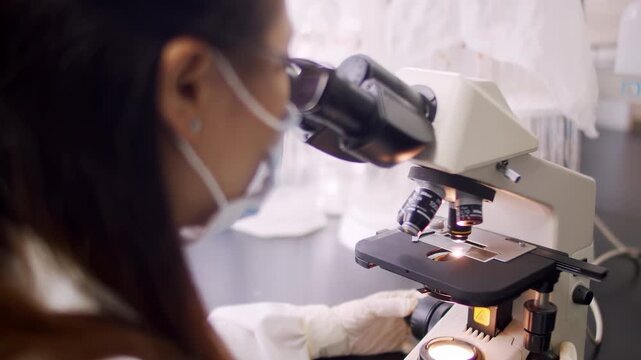 Female scientist in protective suit, goggles and gloves using microscope conducting scientific experiment with petri dishes and test tube it while conducting lab research on antiviral treatment
