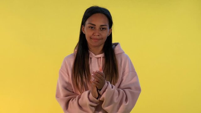 A young black woman with long dark hair wearing a pink hoodie smiles and claps her hands indoors against a solid yellow background in a studio setting.