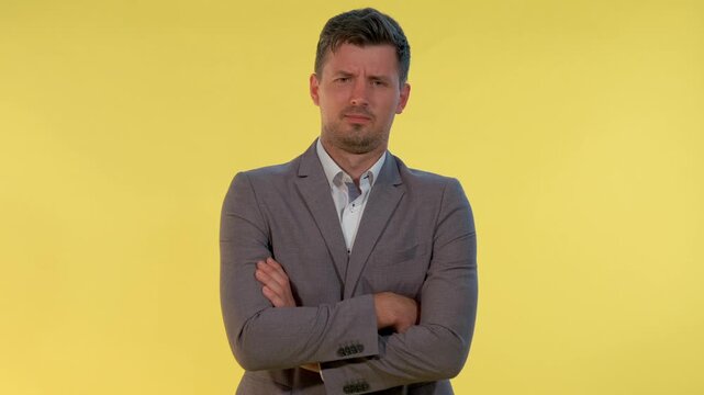 A businessman in a light gray suit jacket gestures with an open hand, asking a question while standing against a solid yellow studio background.