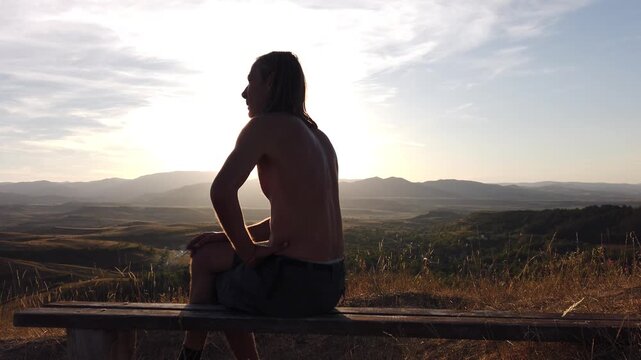 Long haired shirtless man smoking cigarette on mountain bench during sunset