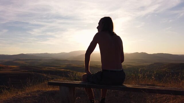 Long haired shirtless man smoking cigarette on mountain bench during sunset