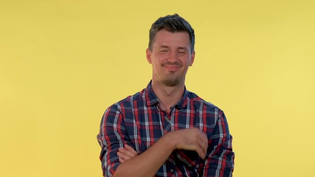 A man in a red, blue, and white plaid shirt poses with crossed arms against a solid yellow background in a bright indoor studio setting.