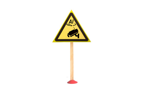 The warning sign features a video camera and an artificial intelligence symbol at the top in the form of a yellow triangle on a light background, captured in a close‑up. The object is under video surv