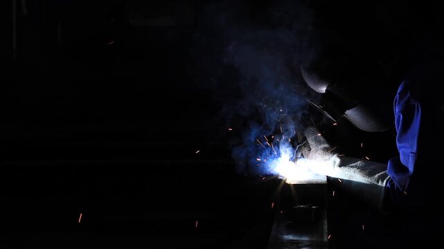 Industrial technician engineer in blue protective cloth wear safety mask, helmet, glasses welding metal. professional welder man welding steel structure with fire spark in factory. selective focus.