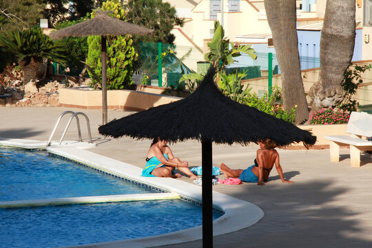 Verano con dos ba&ntilde;istas en la piscina tomando sol en vacaciones con toalla, ba&ntilde;ador y sombrilla de paja pool parasol amigas vacation holidays hablando frente a frente sentadas en el suelo