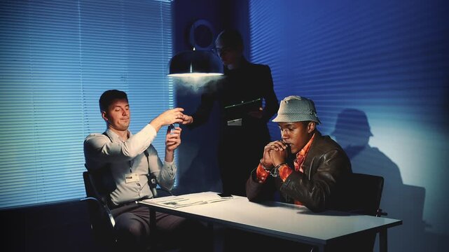 Two men sit across a table in a blue-lit interrogation room while a third figure stands behind them, creating a tense and professional investigative atmosphere.