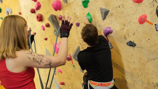 Climber initiates climb on sprayed wall seeking out convenient grips. Female instructor secures safety ropes for sportsman protection