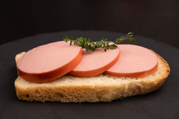 Sausage sandwich background closeup with fresh ham slices on white bread loaf piece, organic pork meat product breakfast appetizer with thyme herb on black slate board, horizontal copy space. © Inna Nikitina