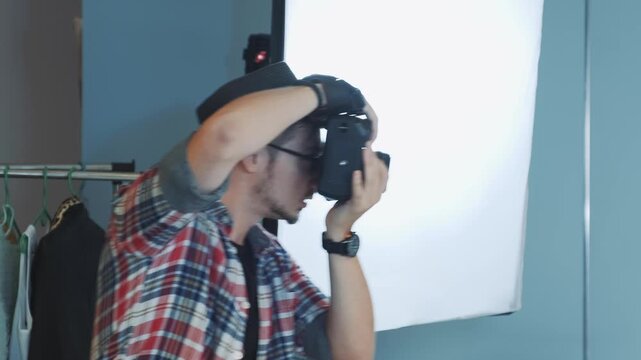 A male photographer wearing a plaid shirt and fedora hat is taking photos with a professional camera in an indoor studio featuring a large softbox and flash unit.