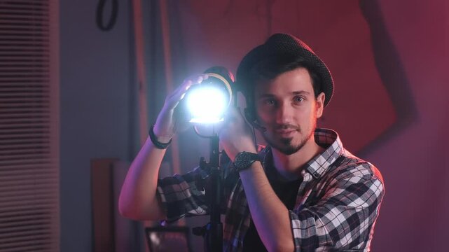 An adult male filmmaker wearing a plaid shirt and fedora adjusts a professional lighting fixture in a studio with red and muted walls.