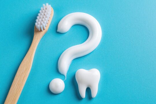 White toothpaste forms question mark shape, tooth icon on blue background. Wooden toothbrush lies nearby. Dental care concept about oral health problems, hygiene questions, choosing right product for