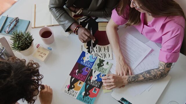 Multiethnic young adult designers analyzing graphic print layouts at office meeting. Footage serves corporate marketing campaigns, branding projects, and collaborative teamwork visuals