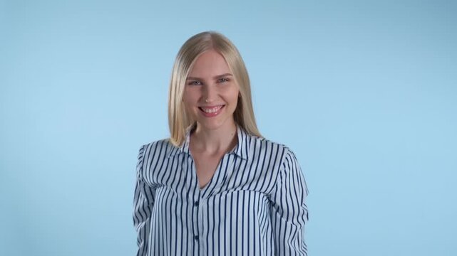 A young blonde woman wearing a blue and white striped shirt slowly turns around and smiles while standing in a studio with a light blue background.