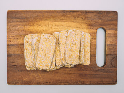 Sliced tempe or tempeh, a traditional Indonesian food made from whole soybeans that are fermented and pressed into a dense, firm cake, on a wooden chopping board with white background