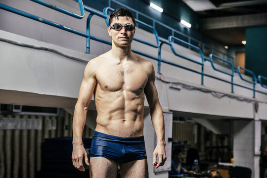 Male swimmer in blue swim trunks and goggles stands confidently at a swimming pool, with bleachers and training equipment visible in the background