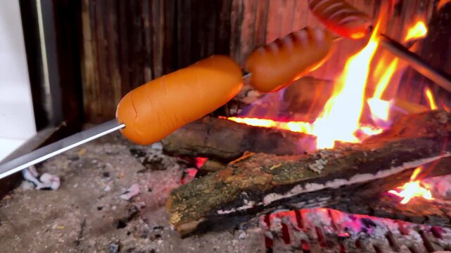 Sizzling sausages cook over bright, crackling flames. Wood burns beneath metal grills, casting warm orange glow. Fire crackles, sizzling meats sizzle, smoke curls upward. Ideal for outdoor cooking