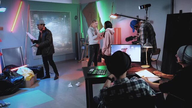 A diverse group of film professionals rehearsing and reviewing documents together in a modern indoor production studio with technical equipment and monitors.