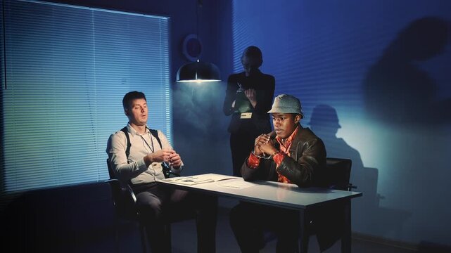 Two men seated at a table in an interrogation room, engaged in a serious discussion with documents spread out, under cool artificial lighting.