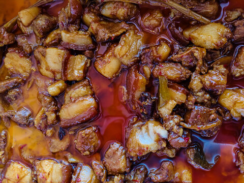 A close-up overhead shot of succulent, braised pork belly, known as 'Babi Kecap'. Tender pieces are bathed in a rich, glossy, dark soy-based sauce with a hint of spice and aromatics.

