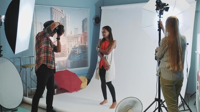 A male photographer takes pictures of two female models posing in a professional indoor photography studio equipped with lighting and camera gear.