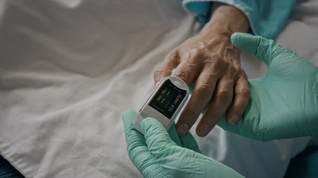 Doctor wearing protective gloves placing pulse oximeter on unrecognizable senior patient finger to check vital signs. Healthcare clip suitable for hospital care campaigns and medical technology ads