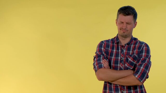 A dark-haired adult man in a red, blue, and white plaid shirt shows negative emotions and gives a thumbs down against a solid yellow background in an indoor studio.