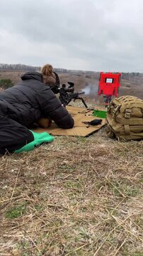 Overcast sky above grassy field with a shooter kneeling. Person in dark coat aims rifle with focused attention. Red equipment box and tan backpack sit nearby. Ideal for training, military