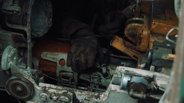 Gloved hands adjusting orange cylinder and coiled spring inside grimy engine compartment with moody low light, worn metal parts, oil staining, and workshop tools nearby