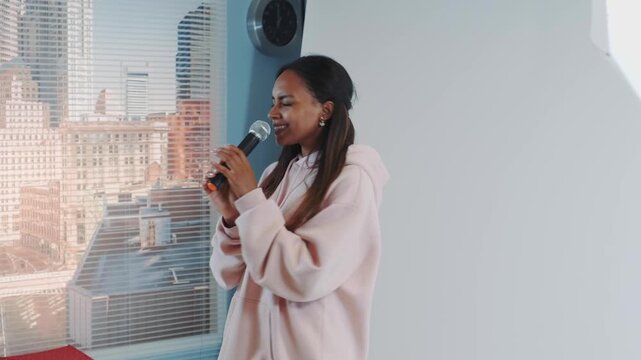 A young woman with dark hair wearing a pink hooded garment sings into a microphone indoors by a large window overlooking a dense urban cityscape during daytime.