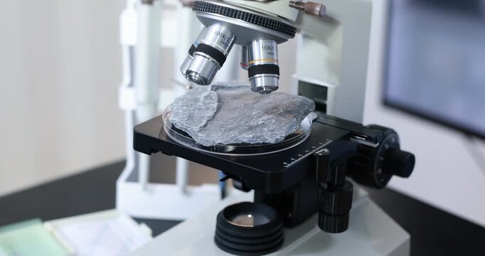 Close-up of a laboratory microscope examining a gray rock sample on a glass slide, scientific research, geology, and material analysis.