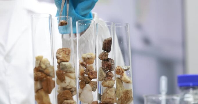 Scientist wearing blue gloves uses tweezers to place mineral rock samples into clear test tubes in a bright laboratory.