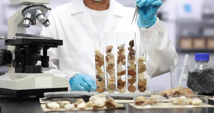 Scientist in a laboratory examines rock and soil samples in glass test tubes beside a microscope, wearing blue protective gloves.