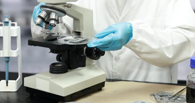 Scientist in a white lab coat and blue gloves adjusts a microscope in a medical research laboratory, with pipettes and glass slides nearby.
