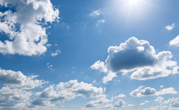 Fluffy white cumulus clouds drift across a vibrant blue sky with bright direct sunlight beaming from the top right corner.