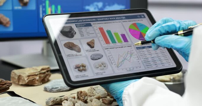 Scientist in blue gloves uses a stylus to study rock and mineral samples with charts on a tablet in a laboratory, with data displayed on a computer screen.