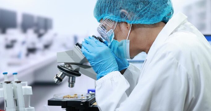 Laboratory researcher in protective clothing examining a sample through a microscope in a modern medical science lab.