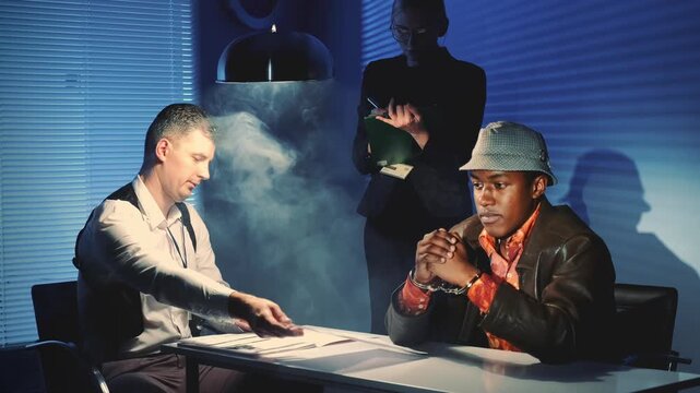 A black prisoner in handcuffs talks with a caucasian detective at a table in an interrogation room while a third person observes in the background.