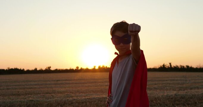 Young superhero boy wears red cape and mask in wheat field at sunset. He raises fist toward camera with confidence. Heroic childhood fantasy inspires courage, imagination, and adventure.