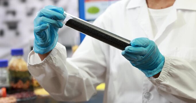 Laboratory scientist in a white coat and blue gloves examines a dark sample tube against a blurred research lab background.