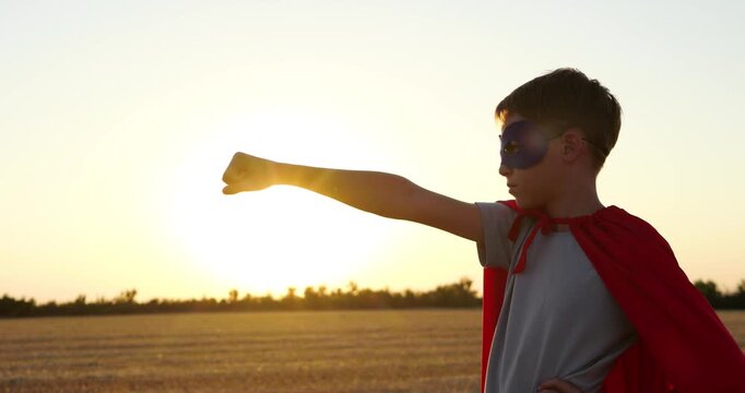 Boy superhero poses in field at sunset. Kid wears red cape and mask, reaching forward like flying hero. Golden sunlight creates inspiring mood for courage, imagination, and childhood dreams.