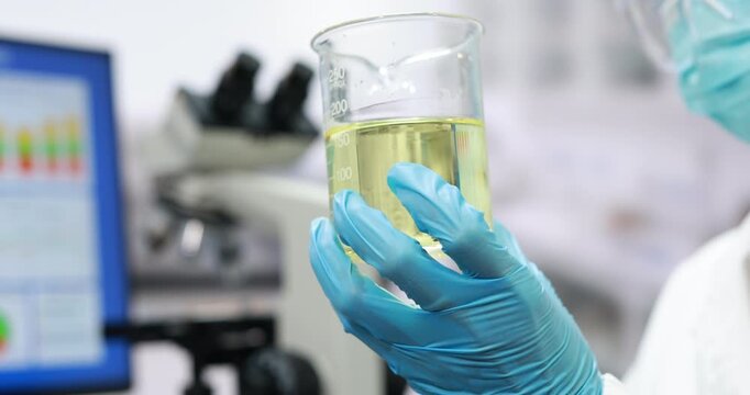 Gloved laboratory researcher holds a glass beaker filled with pale yellow liquid, with a microscope and computer screen blurred in the modern science lab background.