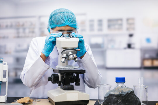 Female scientist in protective gloves and hair cover looks through a microscope in a laboratory, with glassware and rock samples on the workbench.