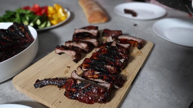 Hand slices smoked ribs on a wooden board. Slices rest beside a bowl of colorful veggies. Knife glints as a person cuts through tender meat. Home kitchen scene suggests comfort and warmth