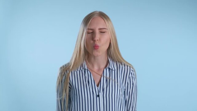 A young blonde woman wearing a blue and white striped shirt makes cheerful and exaggerated funny faces against a solid light blue studio background.