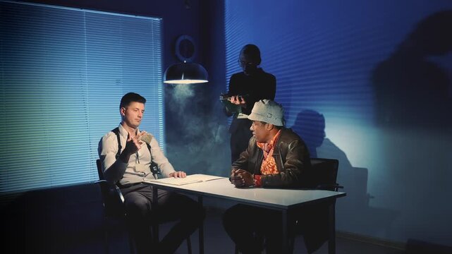Three men engage in a tense discussion around a table in a smoky, industrial-style interrogation room with dramatic lighting and visible documents.