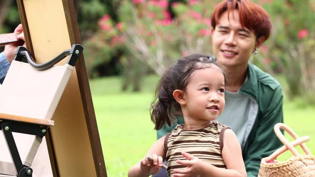 Asian kids painting in park. Asian family enjoy outdoor picnic with food and drinks in park sunny day, outdoor activitie with daughter father, mother grandfather. GrandFather playing acoustic guitar