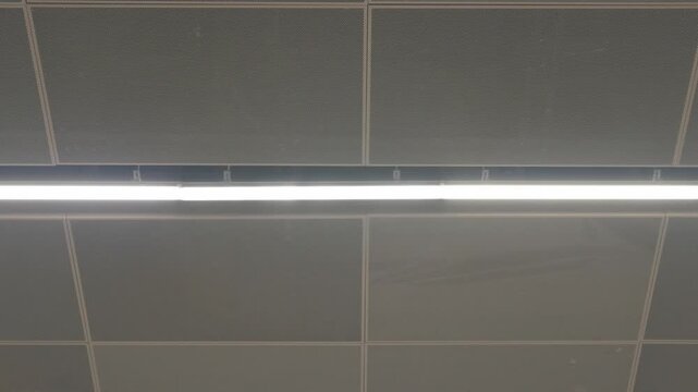 Modern interior ceiling with perforated acoustic panels and linear fluorescent lights, featuring one visibly flashing or flickering light fixture.