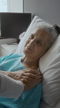 Vertical video of senior Caucasian woman lying in hospital bed receiving touch from caregiver. Clip suits hospice nursing campaigns, geriatric healthcare ads, and palliative care promos